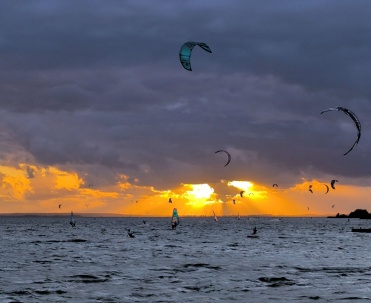 Kitesurferzy na wodzie podczas zachodu słońca &ndash; dynamiczny kadr sportowy z bardzo efektownym niebem.
